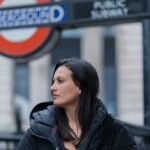 Fashionable woman in black jacket stands by London Underground sign, capturing urban style.