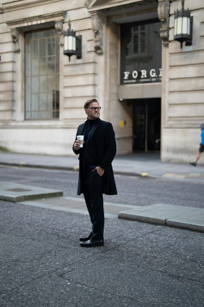 A man in a black suit stands with a coffee cup on a city street, blending modern fashion with urban life.
