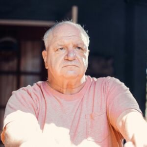 A senior man with a thoughtful expression, sitting outdoors in sunlight.
